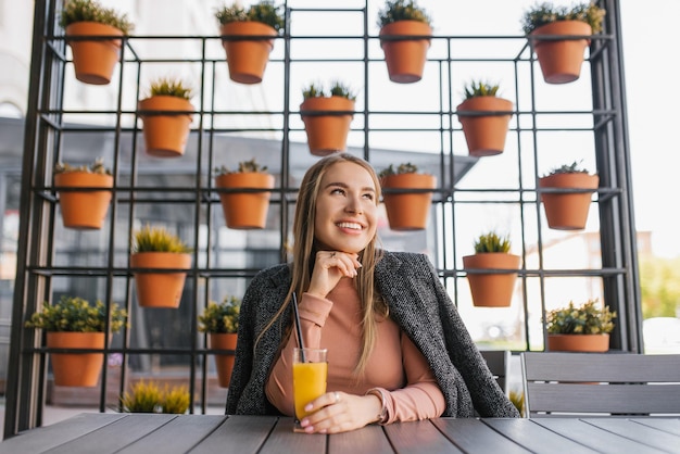 Una joven feliz está sentada en una mesa en un café de verano y bebiendo un vaso de jugo de naranja