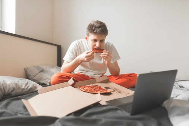 Joven feliz está sentado en la cama con una caja de pizza