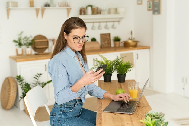 Una joven feliz y hermosa sentada en la mesa en casa trabajando en una computadora portátil sosteniendo un teléfono