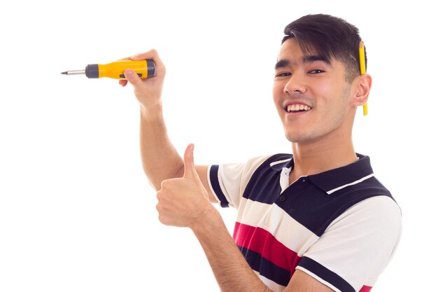 Foto joven feliz con un lápiz detrás de la oreja sosteniendo un destornillador amarillo y mostrando el pulgar hacia arriba