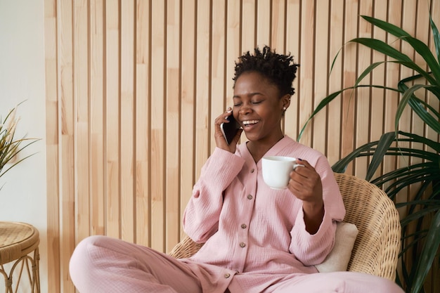 Una joven feliz por la mañana con un acogedor pijama rosa se sienta en una silla de mimbre con una taza de café y