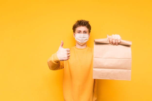 Joven feliz con mascarilla médica aislado de fondo amarillo con bolsa de papel