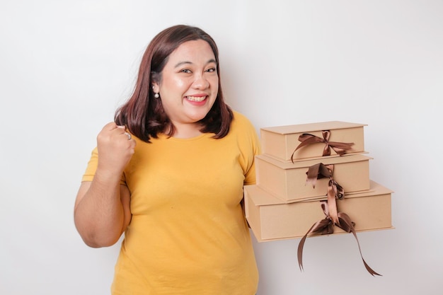 Una joven y feliz mujer asiática de gran tamaño lleva una camisa amarilla y sostiene una caja de regalos y una bolsa de compras