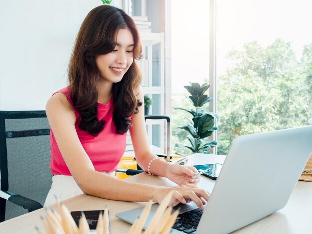 Una joven y feliz mujer asiática que sostiene una tarjeta de crédito mientras usa una computadora portátil para reservar un vuelo en una habitación de hotel para un viaje de verano en casa lista para viajar felices vacaciones concepto de vacaciones de verano