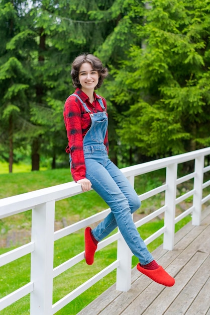 Una joven feliz con overoles de mezclilla azul y camisa roja se sienta en una cerca de madera