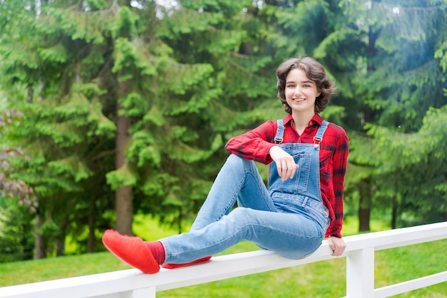 Una joven feliz con overoles de mezclilla azul y camisa roja se sienta en una cerca de madera