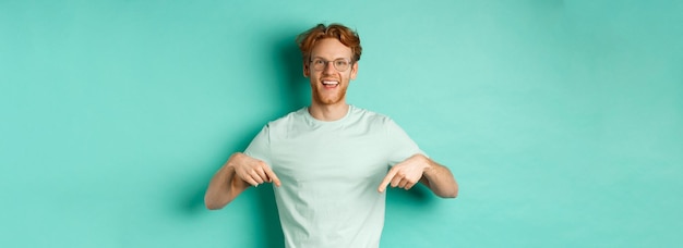 Un joven feliz con el pelo rojo con gafas y una camiseta que muestra un anuncio sonriendo y señalando