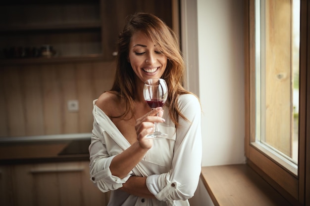 Una joven feliz y pensativa mujer sonriente está bebiendo vino tinto mientras está parada al lado de la ventana.