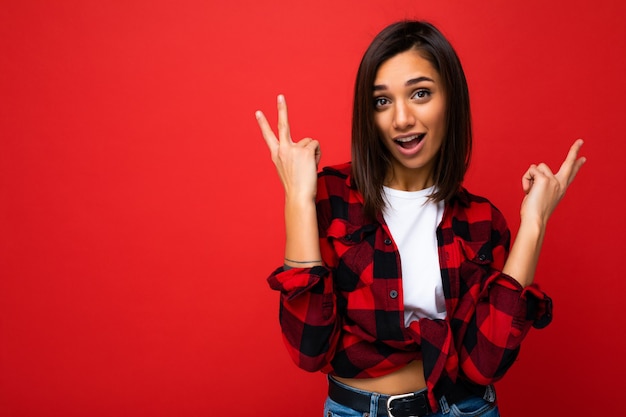 Joven feliz positiva fresca bastante morena con emociones sinceras vistiendo camiseta blanca y camisa a cuadros roja de moda aislada en la pared roja con espacio libre.