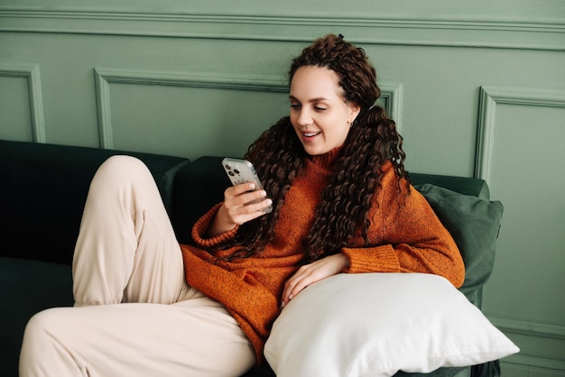 Una joven feliz y relajada sentada en un sofá usando un teléfono celular una mujer sonriente riéndose sosteniendo un teléfono inteligente mirando el teléfono celular disfrutando haciendo compras de comercio electrónico en línea en aplicaciones móviles o viendo videos