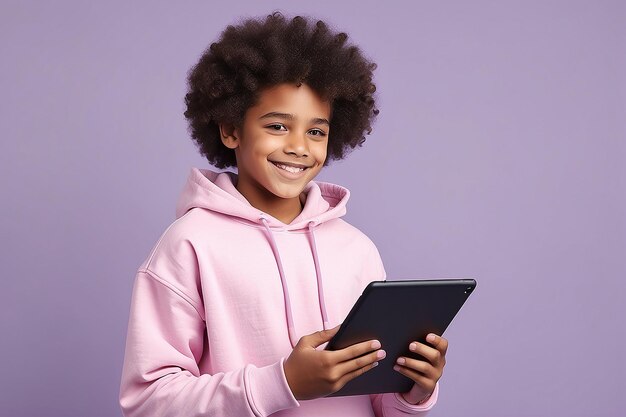Joven feliz y rizado afroamericano adolescente estudiante con sudadera rosada sosteniendo almohadilla usando tecnología de computadora de tableta digital navegando por el aprendizaje electrónico de pie aislado en un fondo púrpura claro