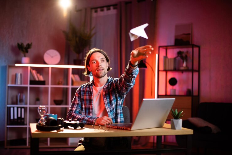 Foto joven feliz con ropa informal tirando un avión de papel mientras toma un descanso durante el trabajo remoto en una laptop