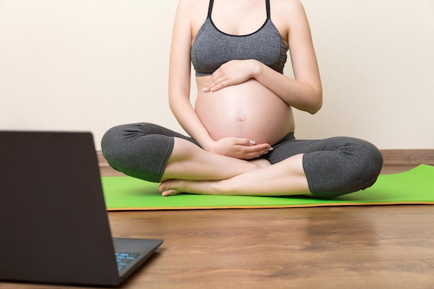 Joven feliz y saludable fitness de yoga hermosa mujer embarazada haciendo ejercicios de yoga bakasana en la estera de yoga frente a la computadora portátil con el concepto de estilo de vida de clase magistral en línea en el momento del coronavirus