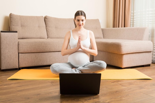 Joven feliz y saludable fitness de yoga hermosa mujer embarazada haciendo ejercicios de yoga bakasana en la estera de yoga frente a la computadora portátil con el concepto de estilo de vida de clase magistral en línea en el momento del coronavirus