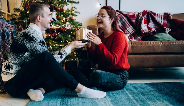 Una joven feliz sentada en el suelo recibiendo un regalo de un hombre en una sala de Navidad decorativa frente al árbol de Navidad celebrando el Año Nuevo en casa mirándose el uno al otro