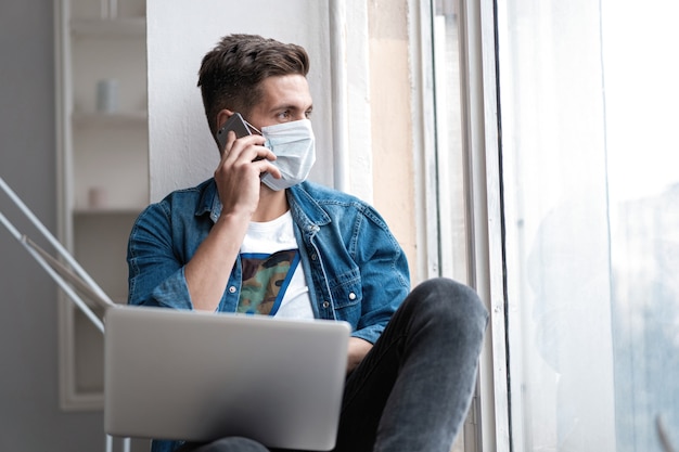 Joven feliz sentado en máscara protectora hablando por teléfono.