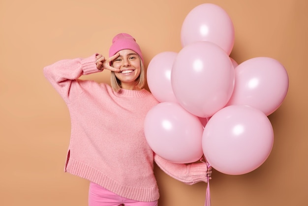 Una joven feliz sonríe ampliamente y hace un gesto de paz sobre los ojos, disfruta de la vida, usa un suéter informal y un sombrero rosa sostiene un montón de globos inflados aislados sobre un fondo marrón Concepto de festividad