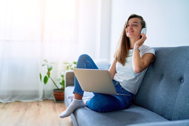 Joven, feliz, sonriente, alegre, alegre, millennial, niña, amante de la música, sentado, en, un, sofá, y, usar la computadora portátil, con, auriculares inalámbricos, para, disfrutar, escuchar, música, en línea, en casa