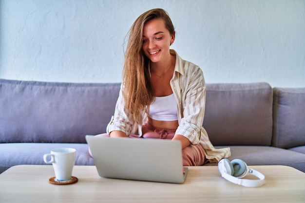 Joven feliz sonriente casual inteligente moderna linda satisfecha chica sentada en el sofá y trabajando en línea en la computadora portátil en casa