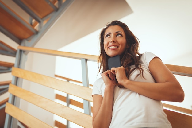 Una joven feliz y sonriente se encuentra en la sala de estar de su nuevo apartamento frente a las escaleras con un teléfono inteligente.