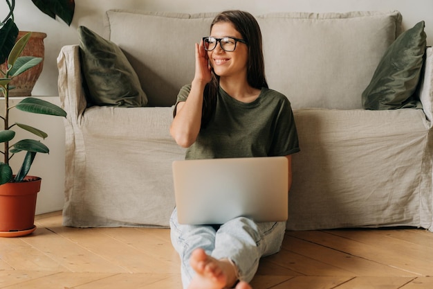 Una joven feliz y sonriente trabajando desde casa en una computadora portátil mientras está sentada en el suelo.