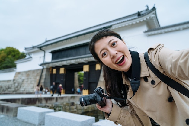 una joven feliz sosteniendo una cámara se está tomando un selfie con un smartphone a la entrada del Castillo de Nijo. una viajera asiática frente a la cámara está emocionada de ver el famoso edificio histórico en Japón.