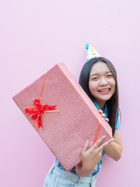 Una joven feliz sostiene una caja de regalos y usa un sombrero de fiesta sobre un fondo rosa