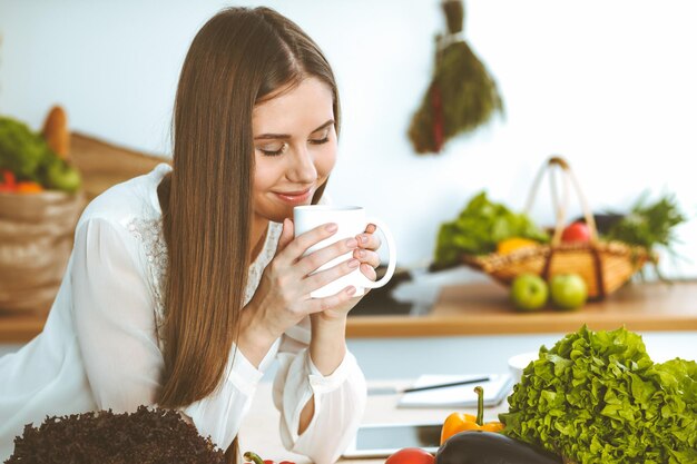 Una joven feliz sostiene una taza blanca y mira la cámara mientras se sienta en una mesa de madera en la cocina entre vegetales verdes