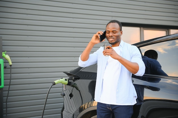 Un joven feliz con un teléfono móvil cargando un coche en una estación de carga de vehículos eléctricos