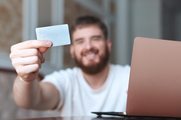 Foto un joven feliz tiene una tarjeta de crédito en la mano, usa una computadora portátil y sonríe en casa comprando en línea