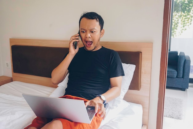 Joven feliz trabajando desde la cama Sostenga la computadora portátil sobre las rodillas y hablando por teléfono