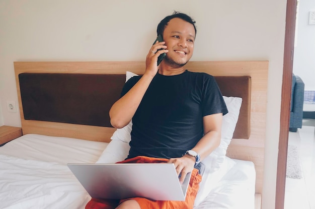 Joven feliz trabajando desde la cama Sostenga la computadora portátil sobre las rodillas y hablando por teléfono