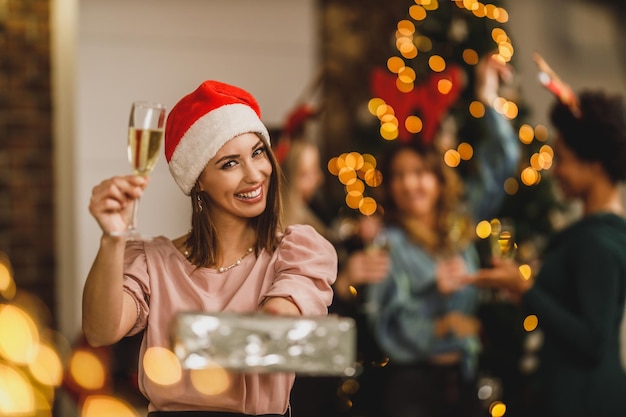 Una joven feliz trayendo regalos y brindando con champán mientras celebra la Navidad con sus amigos en la fiesta de casa.