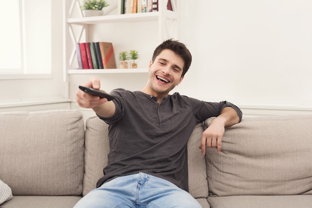 Joven feliz viendo la televisión en el sofá apuntando con el mando a distancia en el televisor, espacio de copia