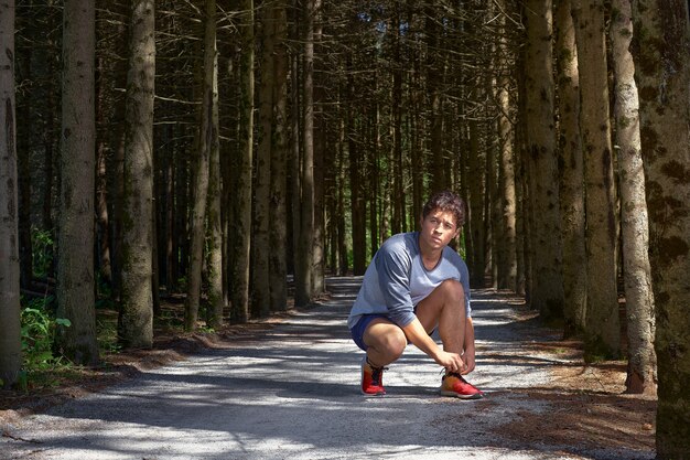 Joven con una figura atlética en pantalones cortos, atando cordones de los zapatos antes de correr por el bosque