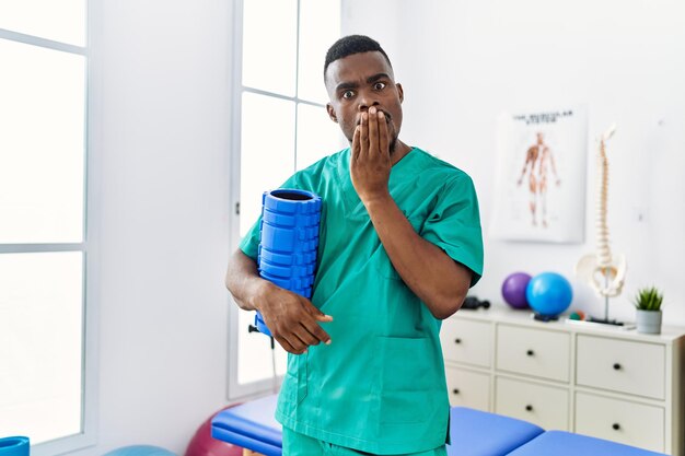 Joven fisioterapeuta africano sosteniendo un rodillo de espuma en la clínica cubriendo la boca con la mano, conmocionado y asustado por un error. expresión sorprendida