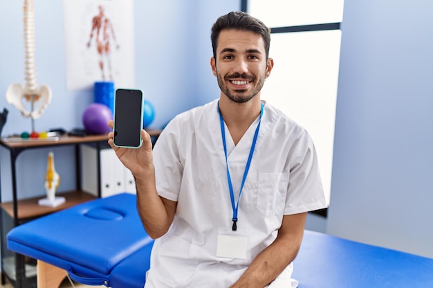 Joven fisioterapeuta hispano sosteniendo un teléfono inteligente en la clínica con un aspecto positivo y feliz de pie y sonriendo con una sonrisa segura mostrando los dientes