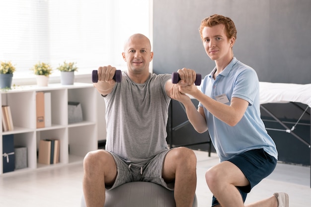 Joven fisioterapeuta y paciente masculino en fitball mirándote durante el ejercicio con pesas en clínicas de rehabilitación o centro médico