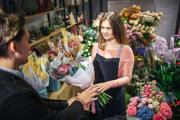 El joven florista de feamel sostiene el ramo y mira al cliente. Ella sonríe. El tipo con chaqueta la mira y alcanza las flores. Se paran adentro.