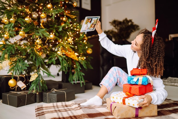 Una joven en el fondo de un árbol de Navidad con regalos con una tableta tiene una videollamada.