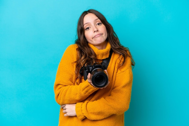 Joven fotógrafa aislada de fondo azul haciendo gestos de duda mientras levanta los hombros