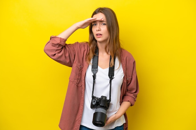 Joven fotógrafa caucásica aislada de fondo amarillo mirando lejos con la mano para mirar algo