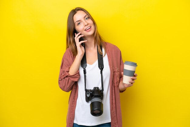 Joven fotógrafa caucásica aislada de fondo amarillo sosteniendo café para llevar y un móvil