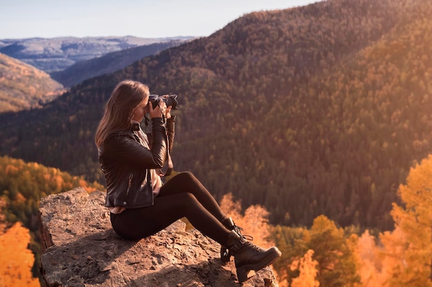 Una joven fotógrafa en un fabuloso paisaje otoñal en las montañas toma fotografías con una cámara profesional sentada en la cima de una montaña rocosa