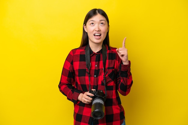 Joven fotógrafa mujer china aislada de fondo amarillo apuntando hacia arriba y sorprendida