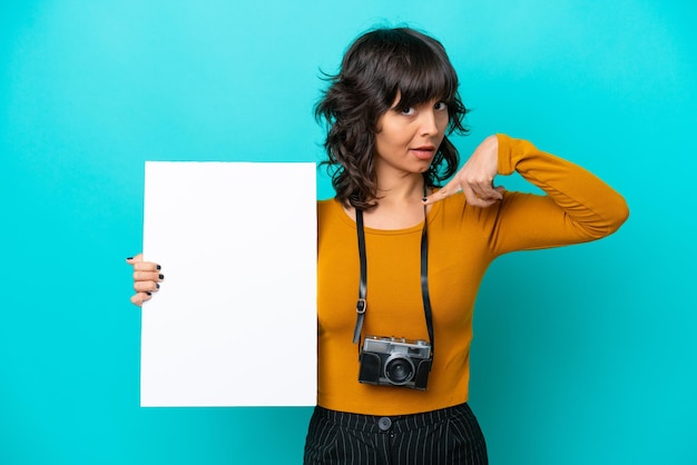 Joven fotógrafa mujer latina aislada de fondo azul sosteniendo un cartel vacío con expresión feliz y apuntándolo