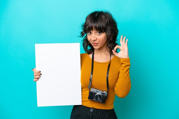 Joven fotógrafa mujer latina aislada de fondo azul sosteniendo un cartel vacío y haciendo el signo de OK