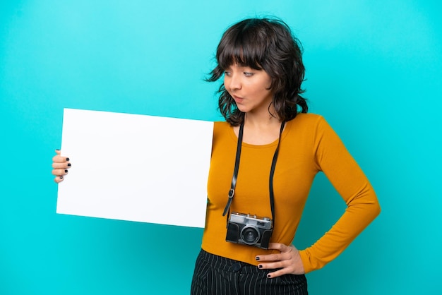 Joven fotógrafa mujer latina aislada de fondo azul sosteniendo un cartel vacío y mirándolo