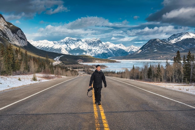 Joven fotógrafo asiático caminando por la carretera con montañas rocosas y lago congelado en invierno