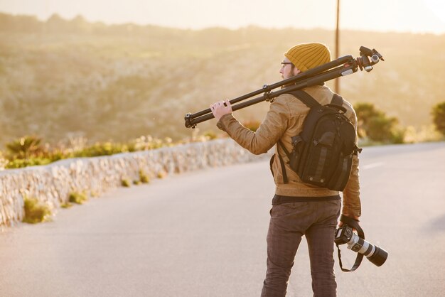 Joven fotógrafo caminando por la carretera del desierto con trípode en su hombro y cámara en mano